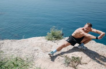 Shirtless man doing a stretch on a cliff overlooking the ocean in daylight.